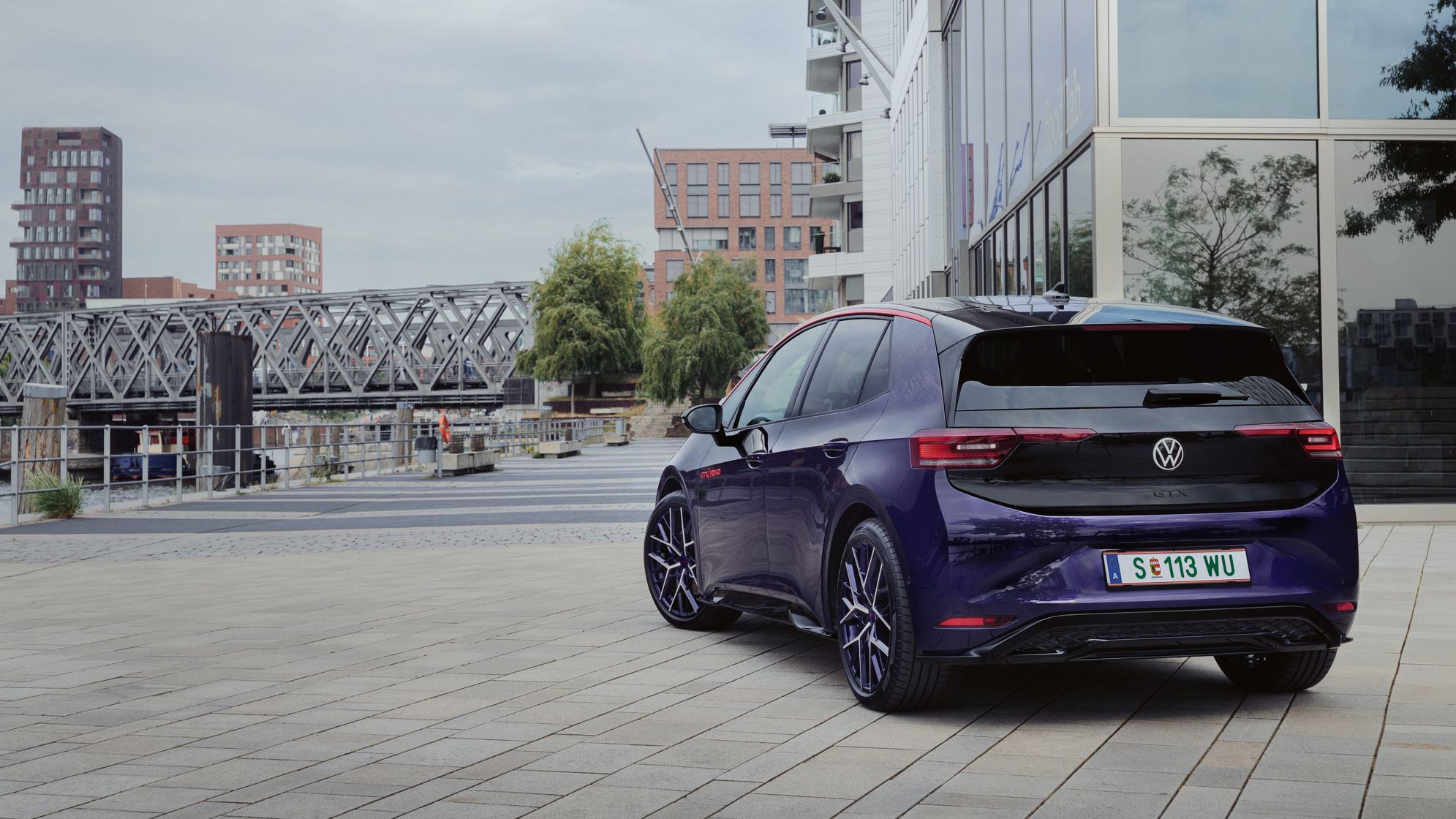 Elektrisches Volkswagen Auto geparkt auf einem modernen Stadtplatz. Im Hintergrund sind Gebäude und eine Brücke zu sehen. Das Auto hat ein Kennzeichen aus Wolfsburg.