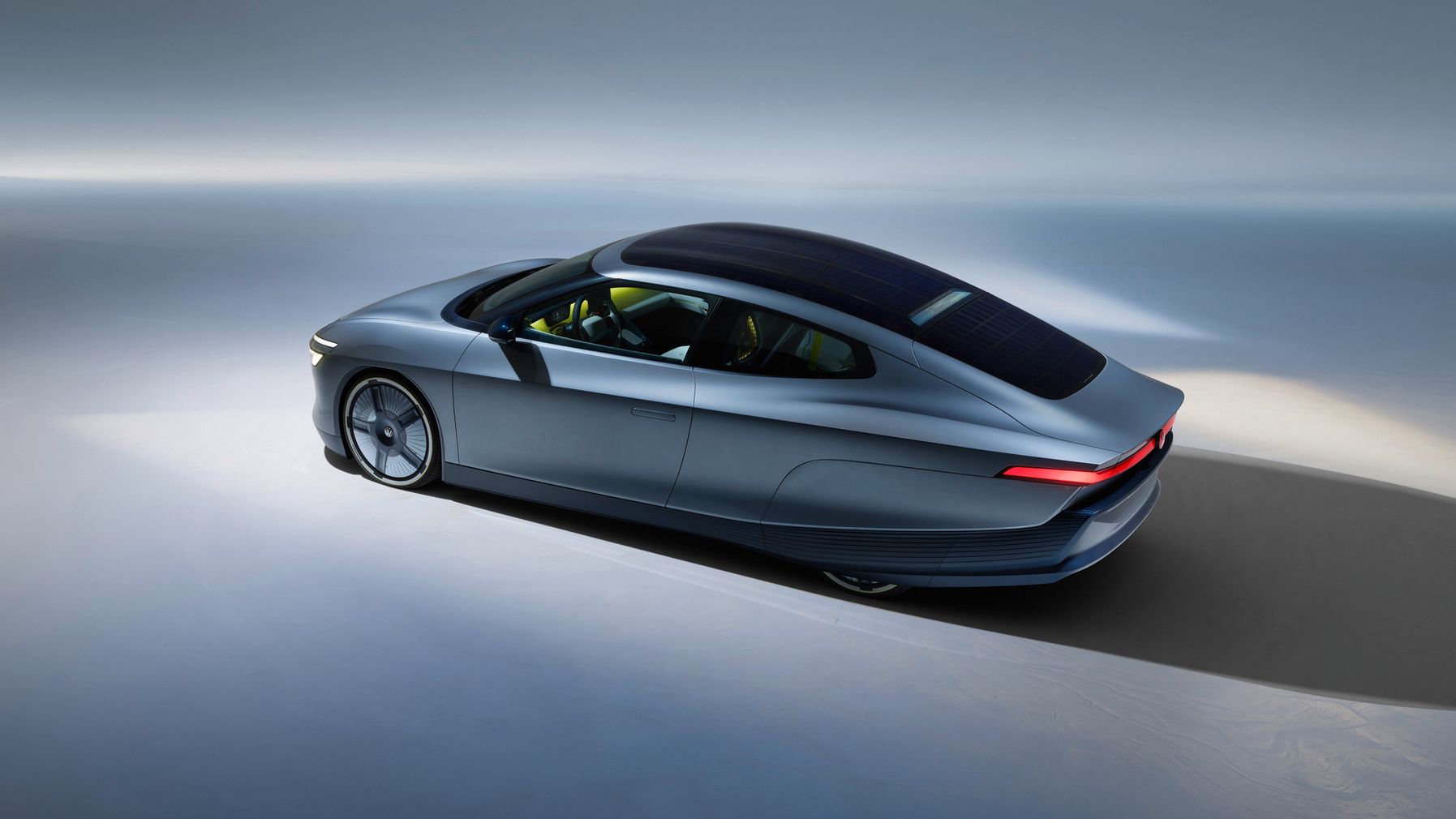 Futuristisches, dunkelgraues Elektroauto mit sehr aerodynamischer, flacher Karosserie fährt in einer minimalistisch ausgeleuchteten Studioumgebung. Das Fahrzeug hat ein glattes, langgezogenes Dach, vermutlich mit integrierten Solarpaneelen, und schmale, leuchtende Rücklichter. Die Szene betont Geschwindigkeit, Effizienz und modernes Design.