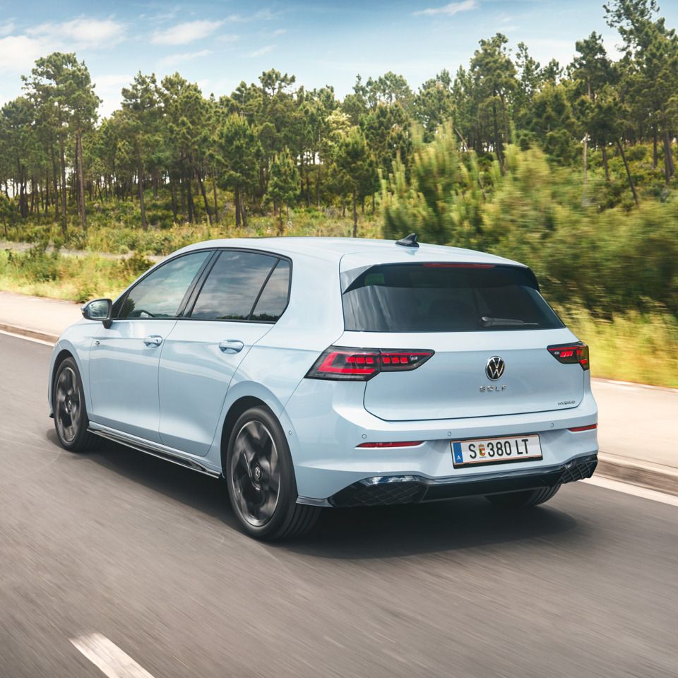 Ein hellblauer VW Golf Hybrid fährt auf einer Straße an einem Wald vorbei.
