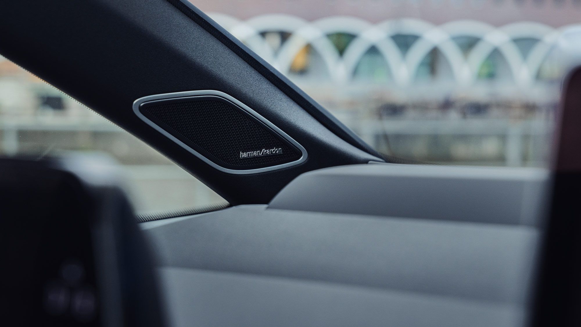 Nahaufnahme des Innenraums eines Autos mit einem Harman Kardon Lautsprecher am Rand des Armaturenbretts. Im Hintergrund sind verschwommen Gebäude und eine gewölbte Struktur zu erkennen. Der Fokus liegt auf dem Lautsprecher und den eleganten Linien des Fahrzeuginnenraums.
