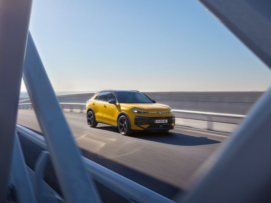 Ein gelber T-Roc fährt über eine moderne Brücke bei klarem Himmel. Die Aufnahme zeigt das Fahrzeug aus der Ferne, teilweise durch die Brückenstrukturen hindurch. Es ist ein sonniger Tag.