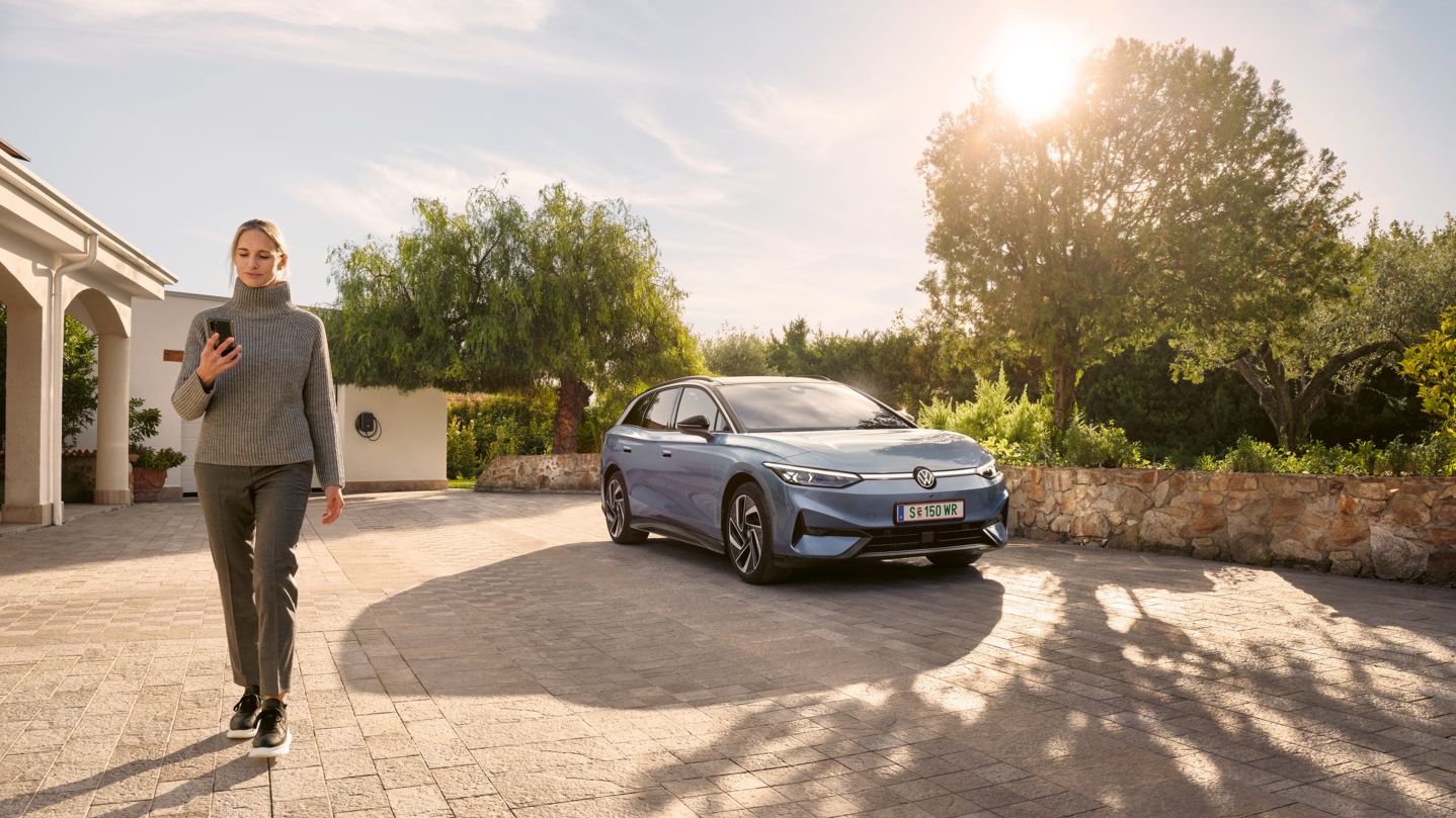 Eine Frau schaut auf ihr Smartphone, im Hintergrund parkt ein blauer VW ID.7 Tourer vor einer Wallbox.