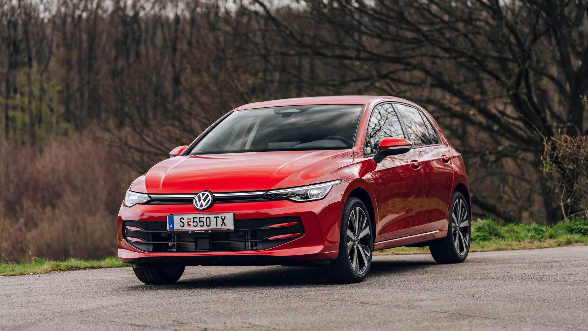 Roter Volkswagen Golf, schräg von vorne fotografiert, steht auf einer Landstraße. Im Hintergrund sind kahle Bäume und eine leicht hügelige, herbstlich wirkende Landschaft zu sehen.