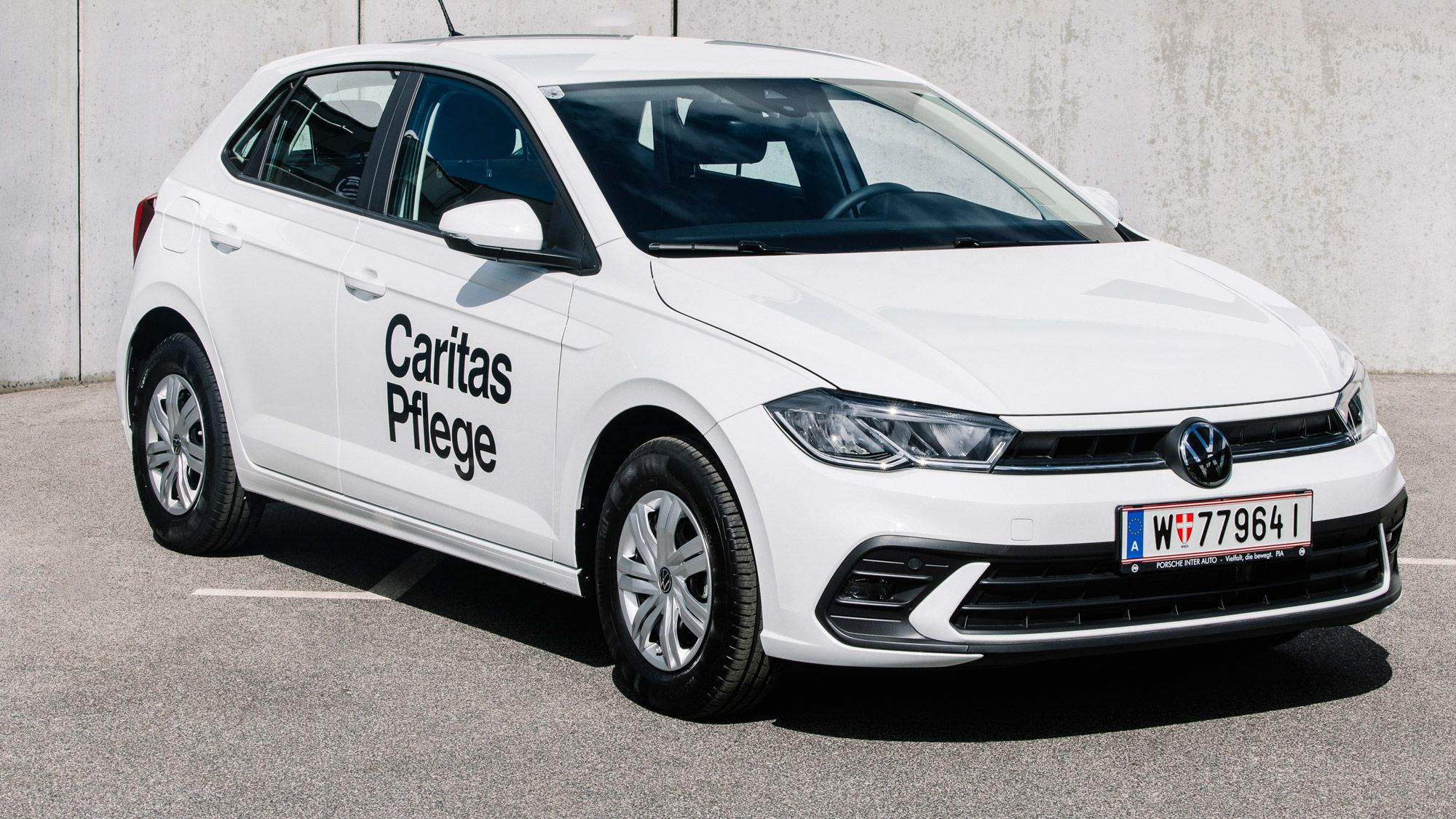 Ein weißer Volkswagen-Polo mit der Aufschrift "Caritas Pflege" auf der Tür steht auf einem Parkplatz vor einer Betonwand. Das Auto hat ein österreichisches Kennzeichen. Die Umgebung ist spärlich und besteht hauptsächlich aus Asphalt und Beton.