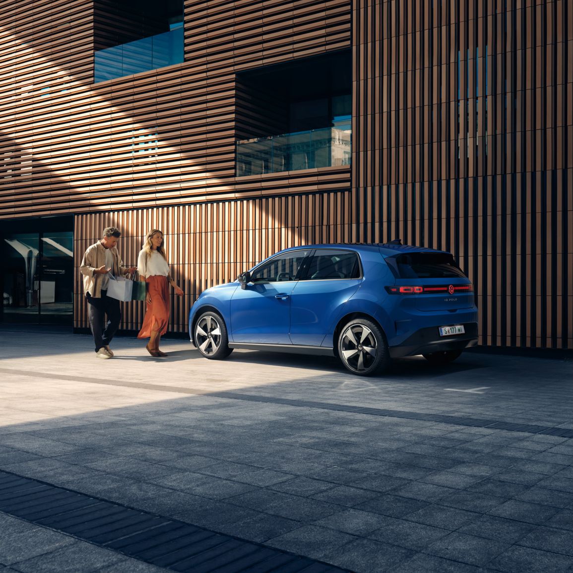 Ein blauer VW ID. Polo Style steht vor einem Gebäude mit Holzfassade. Ein Mann und eine Frau mit Einkaufstaschen gehen darauf zu