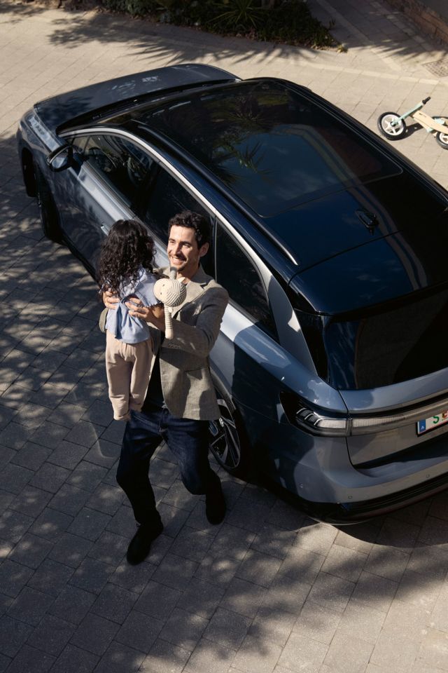 Mann hebt sein Kind hoch, was eine Puppe in der Hand halt und dahinter steht geparkt in der Einfahrt ein ID.7 Tourer und ein umgekipptes Kinderfahrrad.