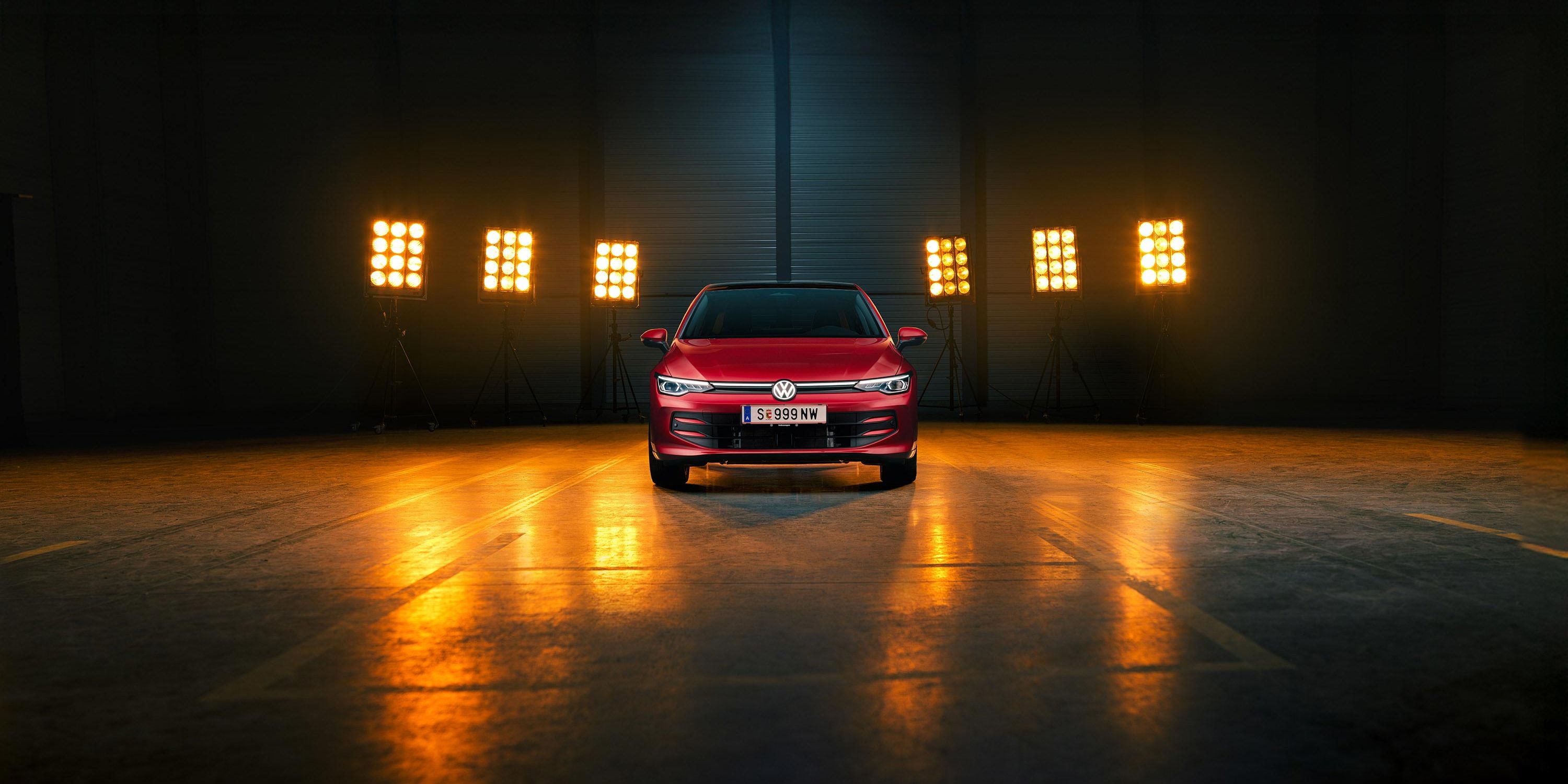 Ein roter VW Golf Falco Edition in Frontalansicht steht mittig im Bild in einer leeren Halle, Bühnenlichter strahlen das Fahrzeug von hinten an.