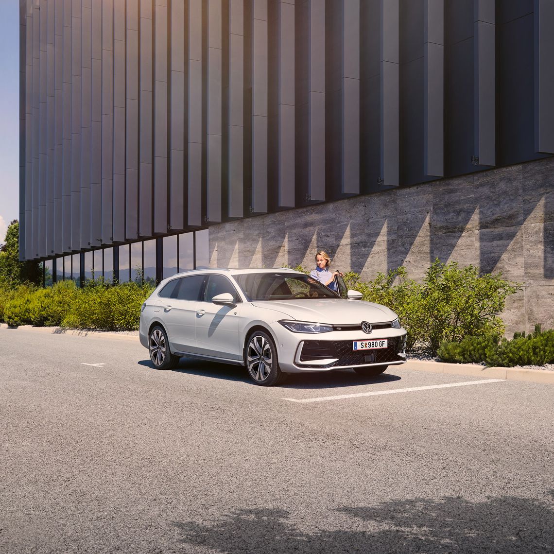  Eine Frau öffnet die Fahrertür eines weißen VW Passat R-Line und steigt ein. Im Hintergrund sieht man ein Gebäude.