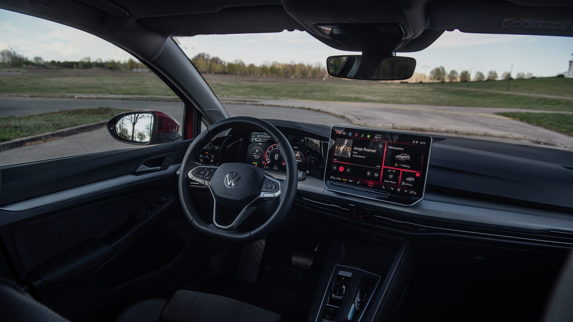 Innenraum eines modernen VW-PKW mit Fokus auf Cockpit und Armaturenbrett, aufgenommen bei Tageslicht. Vor dem Fahrersitz befindet sich ein digitales Kombiinstrument, daneben ein großer Touchscreen mit Infotainment-Anzeige. Lenkrad und Mittelkonsole wirken sportlich und minimalistisch, im Hintergrund ist durch die Scheibe eine grüne Wiese zu sehen.