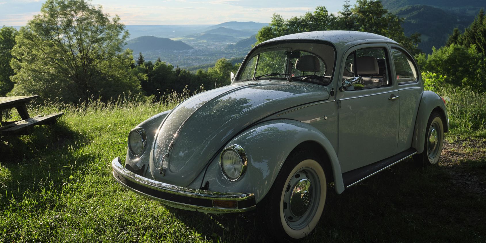 Ein grauer VW Käfer steht auf einer grünen Wiese. Im Hintergrund sind Bäume und hügelige Landschaften unter einem bewölkten Himmel zu sehen. Die Sonne scheint durch die Wolken und beleuchtet das Auto.