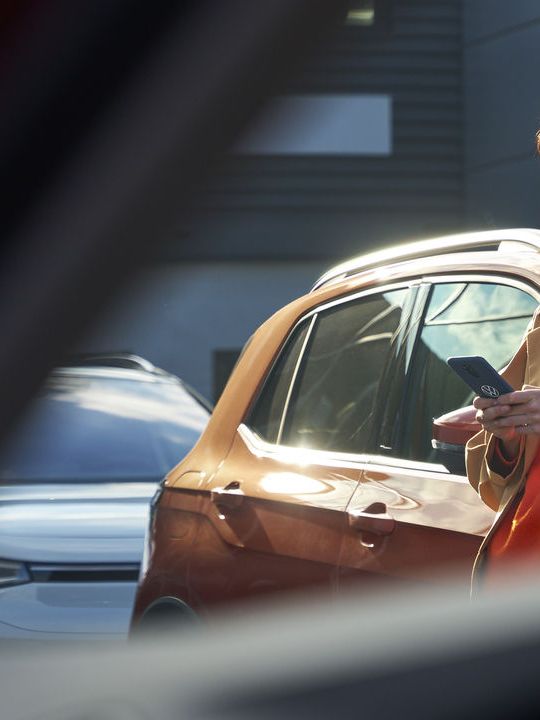 Eine Frau lehnt an einem Volkswagen und blickt  auf ihr Smartphone.