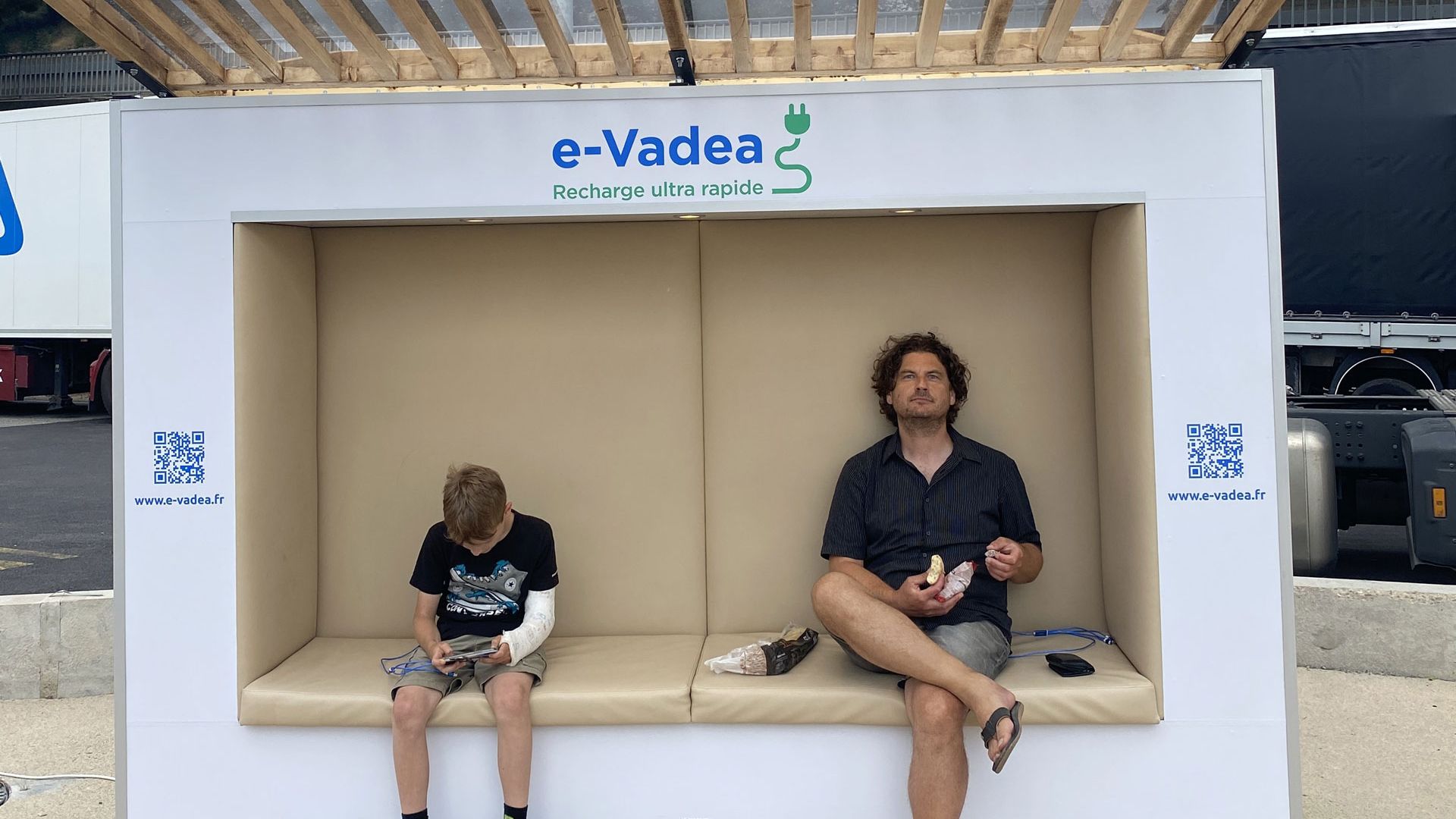 Ein Mann und ein Junge sitzen auf einer Ladebank für Elektroautos. Der Mann isst ein Sandwich, während der Junge auf sein Handy schaut. Beide entspannen sich unter einem schattenspendenden Dach.