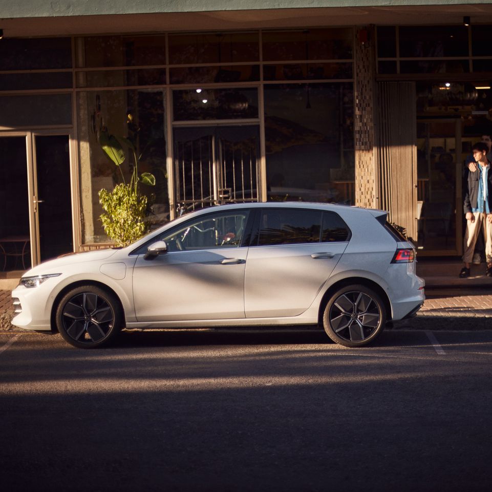 Ein weißer Golf eHybrid parkt am Straßenrand vor einem Café. 