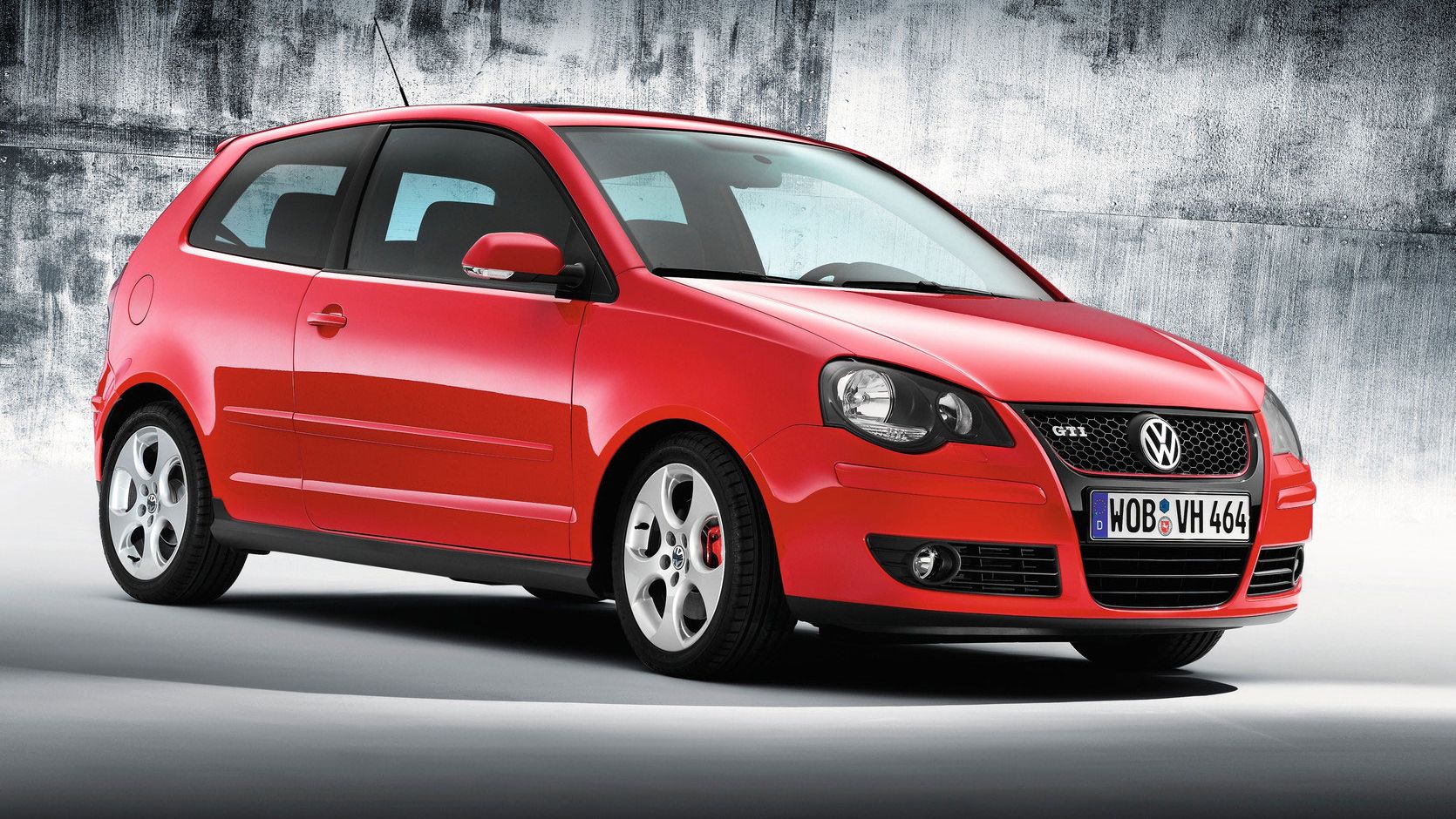 Das Bild zeigt einen roten VW Polo 4 GTI vor einem grauen, strukturierten Hintergrund. Der Wagen ist seitlich und von vorne zu sehen. Die Kennzeichen zeigen „WOB-VH 464“.