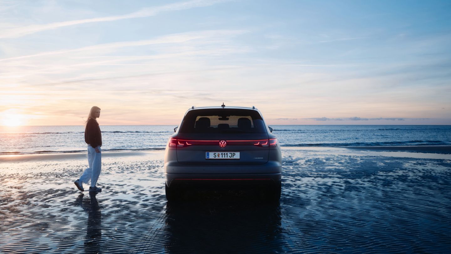 Ein Touareg R steht am Strand bei Sonnenuntergang. Eine Person spaziert nahe beim Fahrzeug. Der Himmel ist klar mit wenigen Wolken.