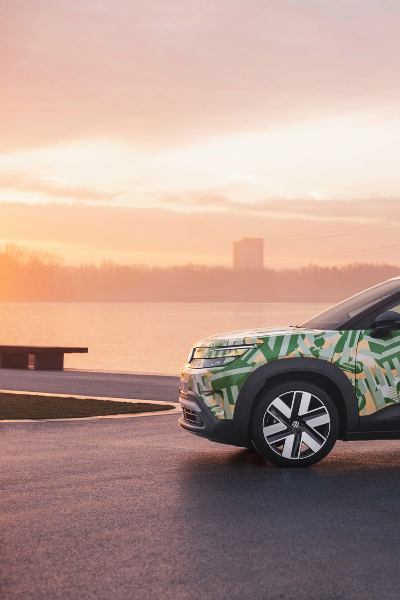 Ein SUV mit einer grünen und weißen Tarnlackierung steht auf einem asphaltierten Platz vor einem Gewässer bei Sonnenuntergang. Im Hintergrund sind Strommasten und Gebäude zu sehen. Der Himmel ist leicht bewölkt und in warmen Farben gehalten.