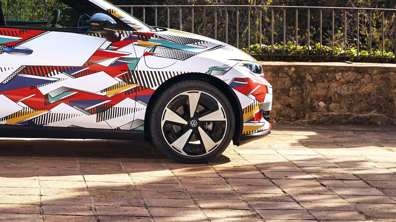 Nahaufnahme eines ID. Polo mit buntem geometrischem Design und einem Volkswagen-Logo auf der Felge. Das Auto steht auf einem gepflasterten Weg, im Hintergrund ist eine niedrige Steinmauer und eine grüne Landschaft zu sehen.