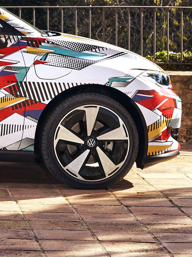 Nahaufnahme eines ID. Polo mit buntem geometrischem Design und einem Volkswagen-Logo auf der Felge. Das Auto steht auf einem gepflasterten Weg, im Hintergrund ist eine niedrige Steinmauer und eine grüne Landschaft zu sehen.