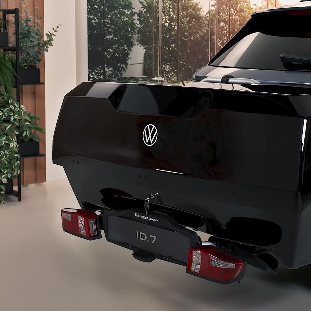 Das Bild zeigt die Rückseite eines schwarzen Volkswagen ID.7 in einem Innenraum. Die Rücklichter sind deutlich sichtbar und es sind Pflanzen im Hintergrund zu erkennen.