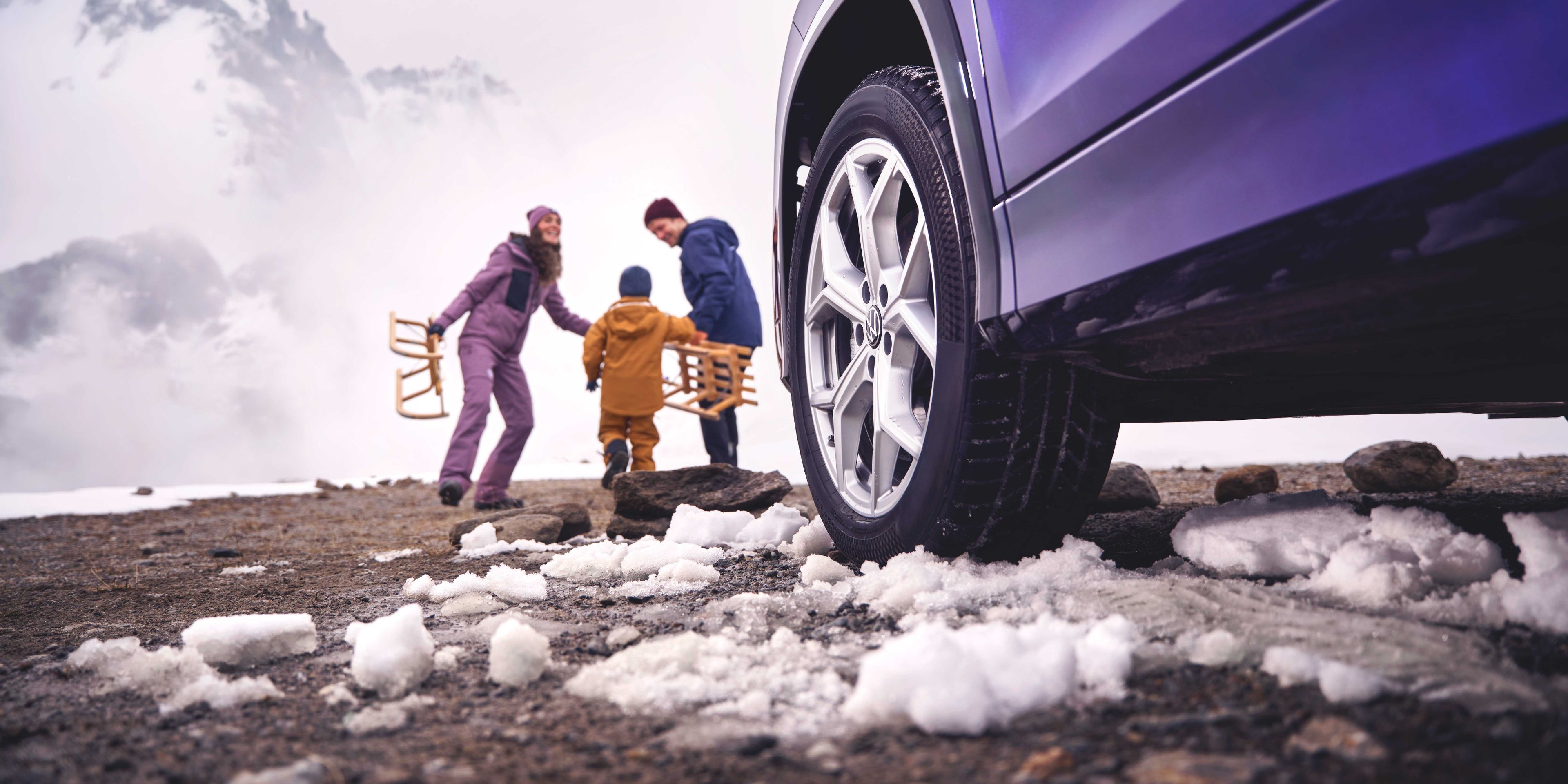 Blick vorbei an der linken Frontseite eines Volkswagen Tayron auf eine Familie mit Schlitten in einer verschneiten Berglandschaft.