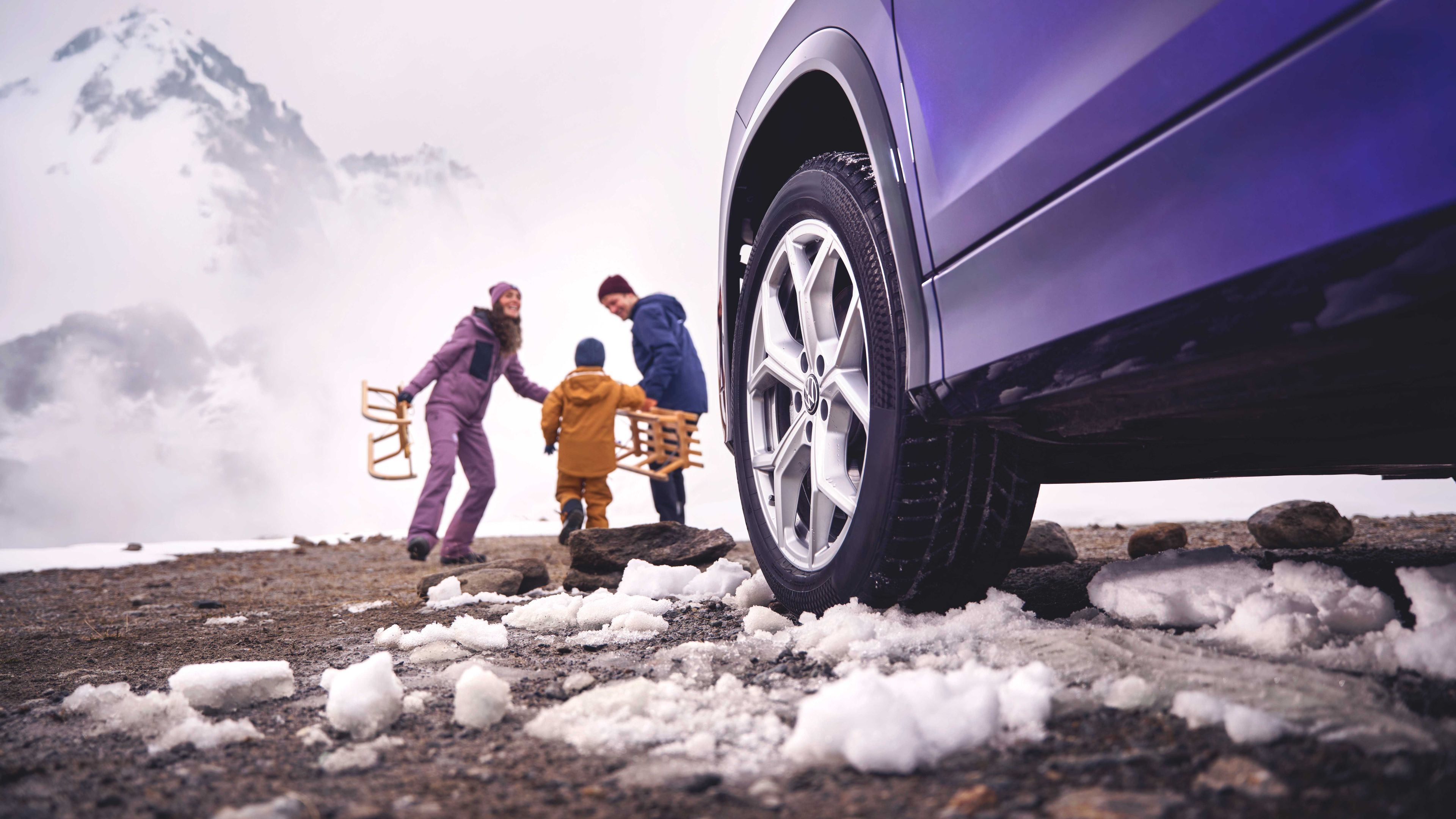 Blick vorbei an der linken Frontseite eines Volkswagen Tayron auf eine Familie mit Schlitten in einer verschneiten Berglandschaft.