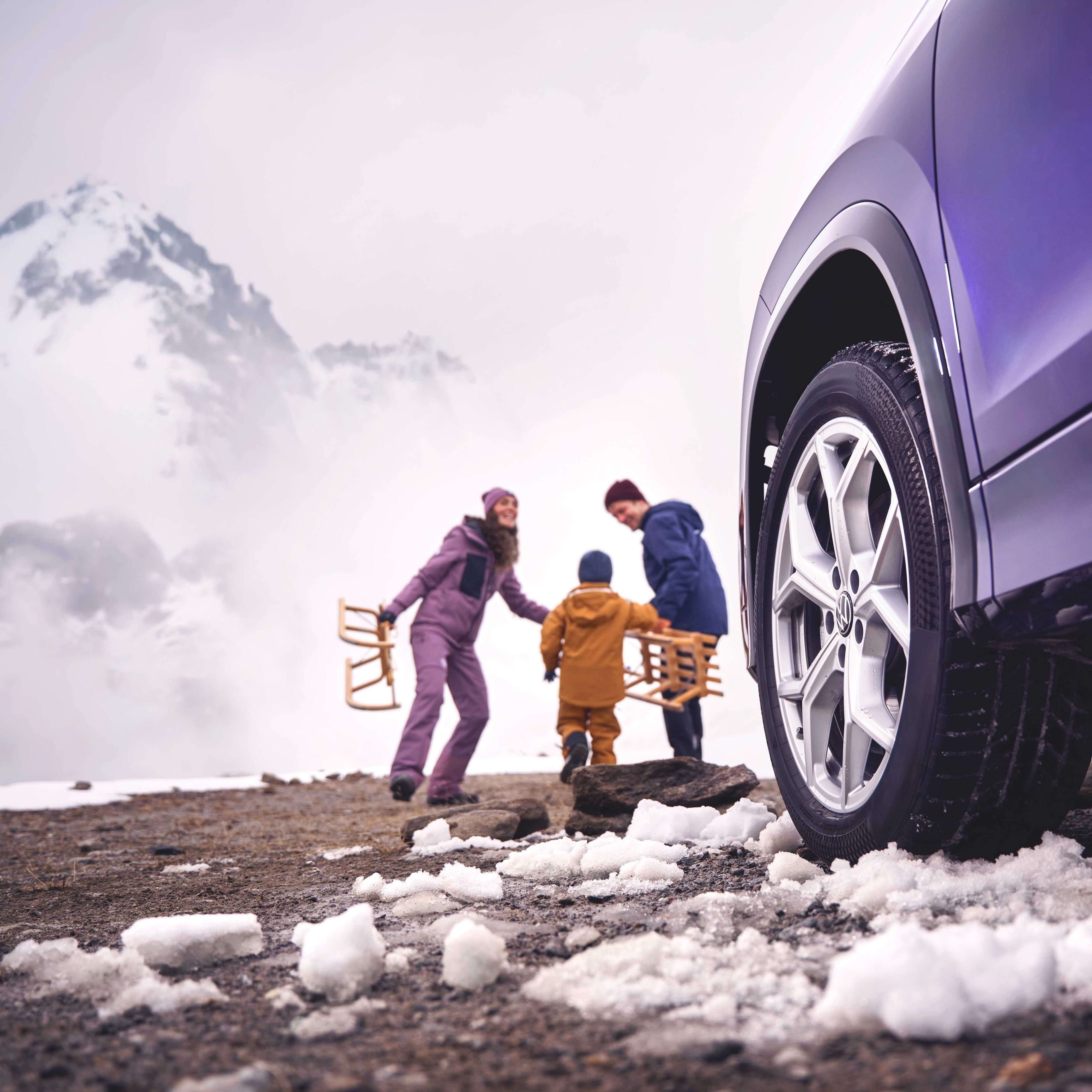 Blick vorbei an der linken Frontseite eines Volkswagen Tayron auf eine Familie mit Schlitten in einer verschneiten Berglandschaft.