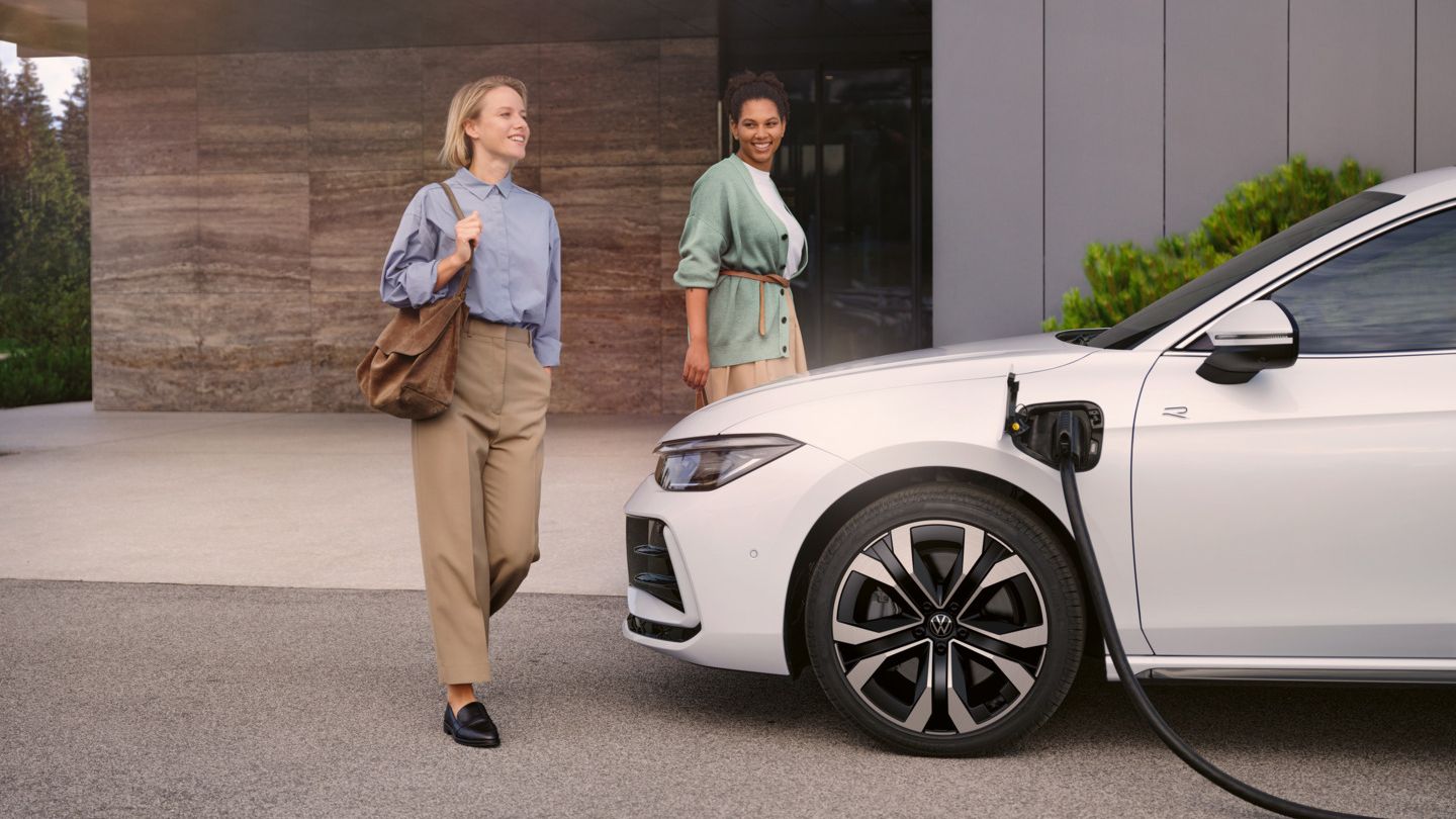 Zwei Frauen gehen auf einen weißen VW Passat R-Line eHybrid zu, der gerade geladen wird.