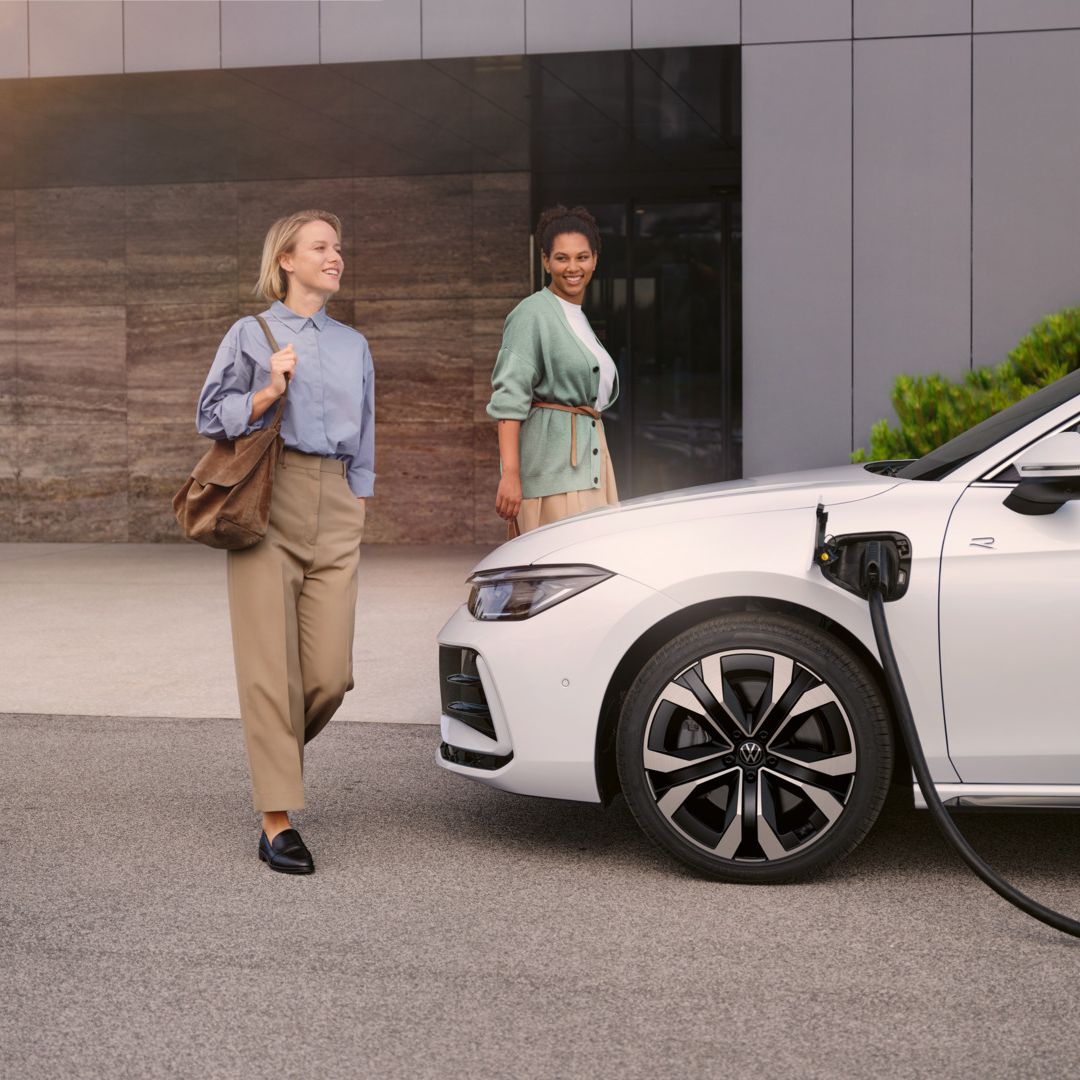 Zwei Frauen gehen auf einen weißen VW Passat R-Line eHybrid zu, der gerade geladen wird.