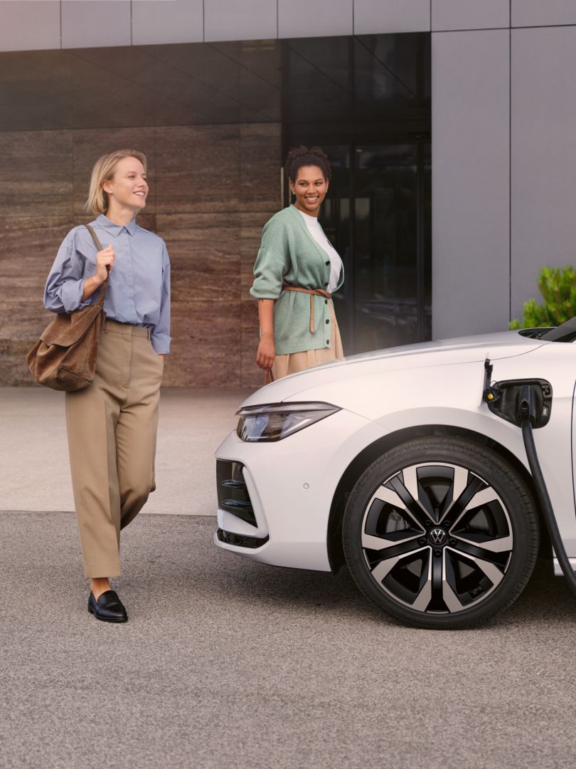Zwei Frauen gehen auf einen weißen VW Passat R-Line eHybrid zu, der gerade geladen wird.