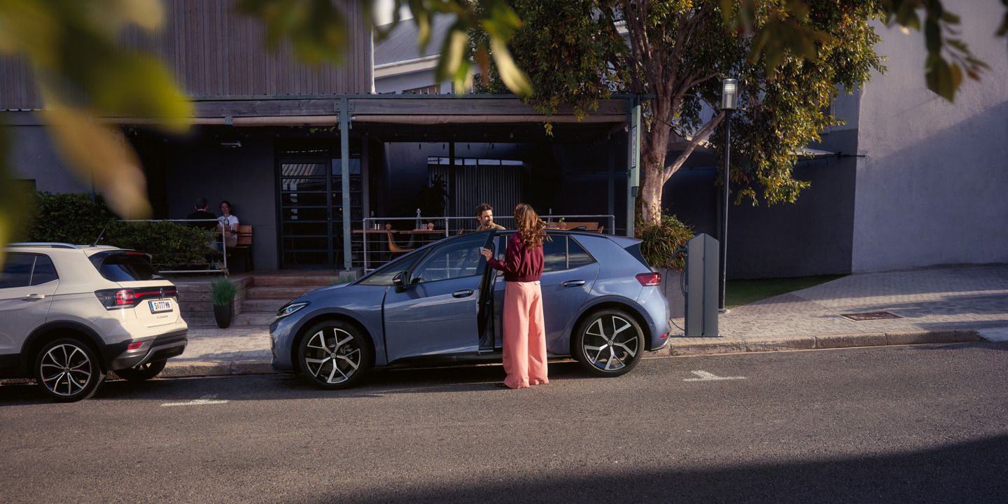 Eine Frau und ein Mann steigen in einen blauen VW ID.3 Neo, der vor einem Café parkt