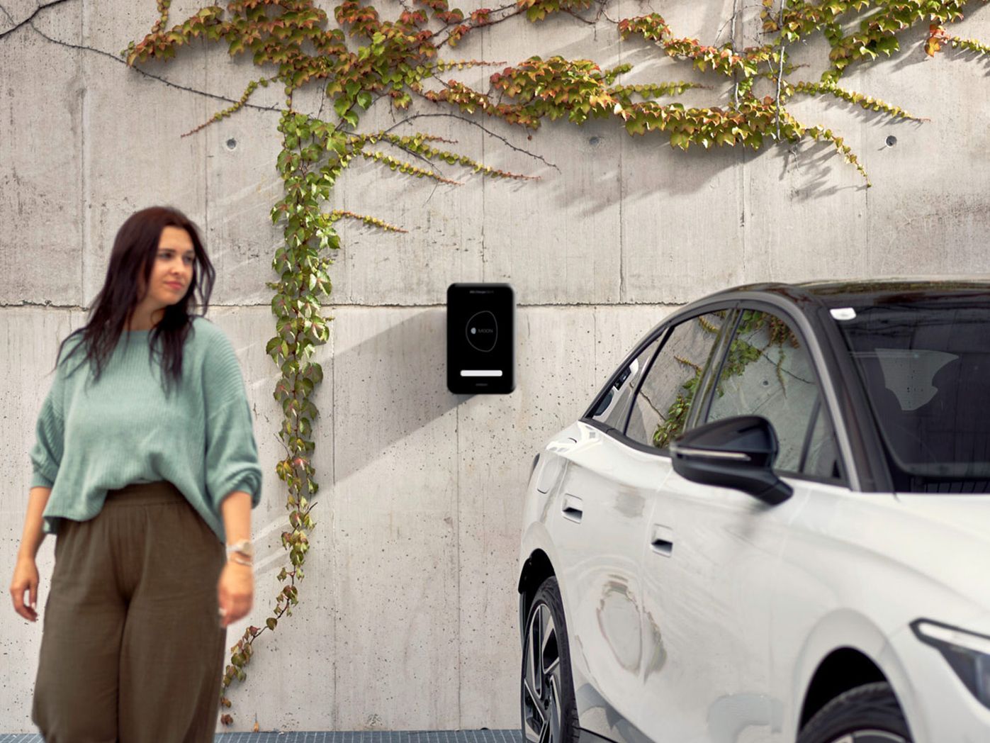 Eine Frau geht in der Nähe eines weißen ID.7, das an einer Wand mit einer Ladestation und Kletterpflanzen geparkt ist. Die Umgebung wirkt urban und modern.