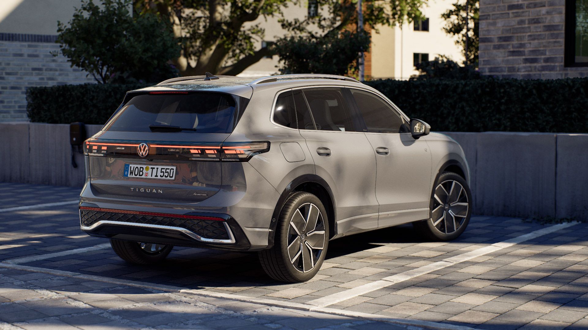 Ein silbergrauer VW Tiguan steht auf einem gepflasterten Parkplatz neben einem modernen Gebäude. Das Fahrzeug ist von hinten schräg zu sehen, mit markanten LED-Rückleuchten und sportlichen Felgen. Im Hintergrund sind eine niedrige Mauer, Hecken und Bäume zu erkennen.