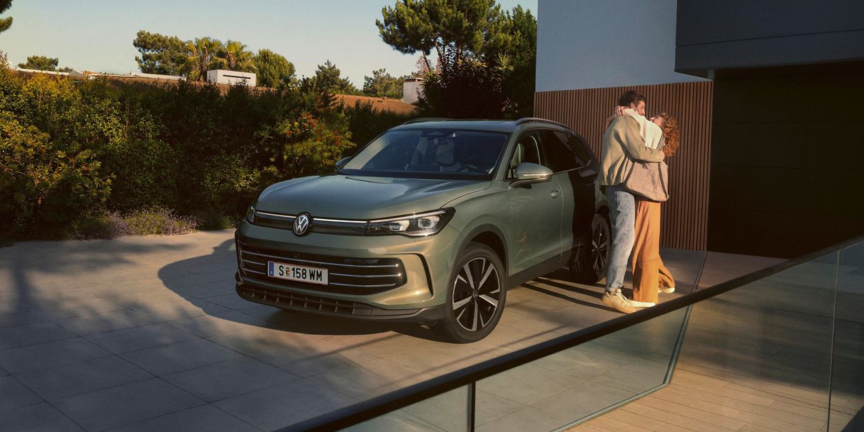 Eine Frau und ein Mann umarmen sich, neben ihnen steht ein VW Tiguan Elegance in der Farbe Cipressino-Grün Metallic