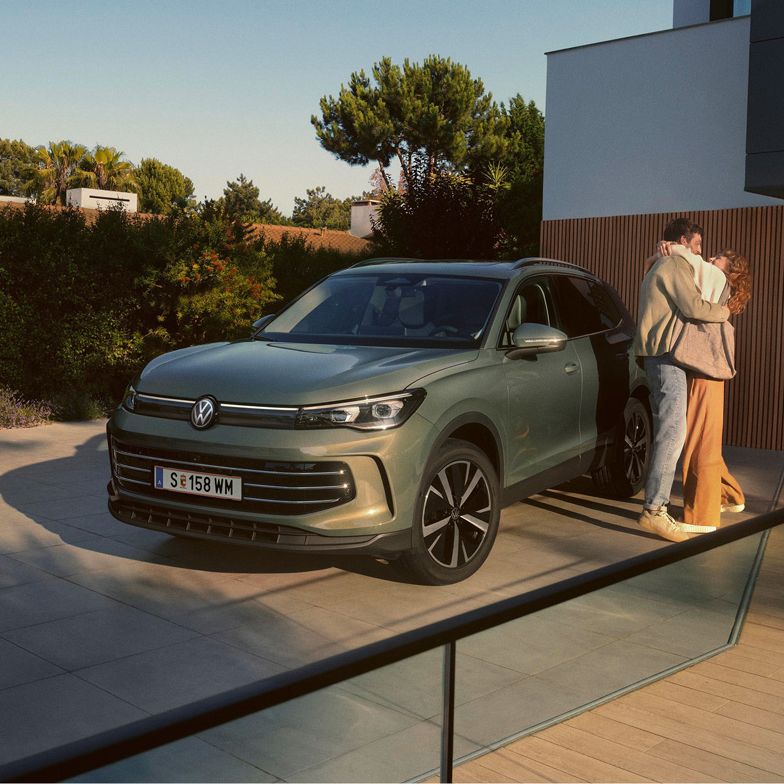 Eine Frau und ein Mann umarmen sich, neben ihnen steht ein VW Tiguan Elegance in der Farbe Cipressino-Grün Metallic