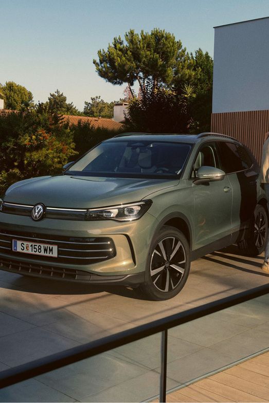 Eine Frau und ein Mann umarmen sich, neben ihnen steht ein VW Tiguan Elegance in der Farbe Cipressino-Grün Metallic
