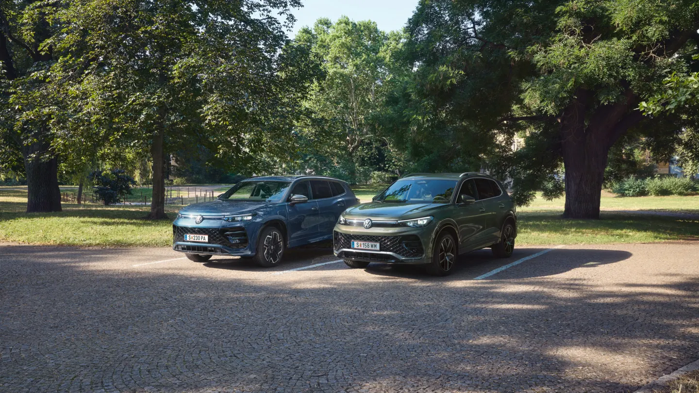 Blauer Tiguan R-Line Plug-In-Hybrid und grüner Tayron R-Line Plug-In- Hybrid parken nebeneinander im schattigen Parkbereich.