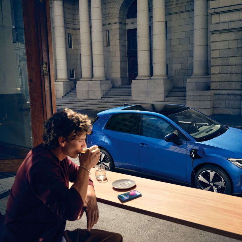 Ein Mann sitzt in einem Café, durch ein offenes Fenster ist der blaue VW ID. Polo Style zu sehen, der an einer Ladestation lädt.