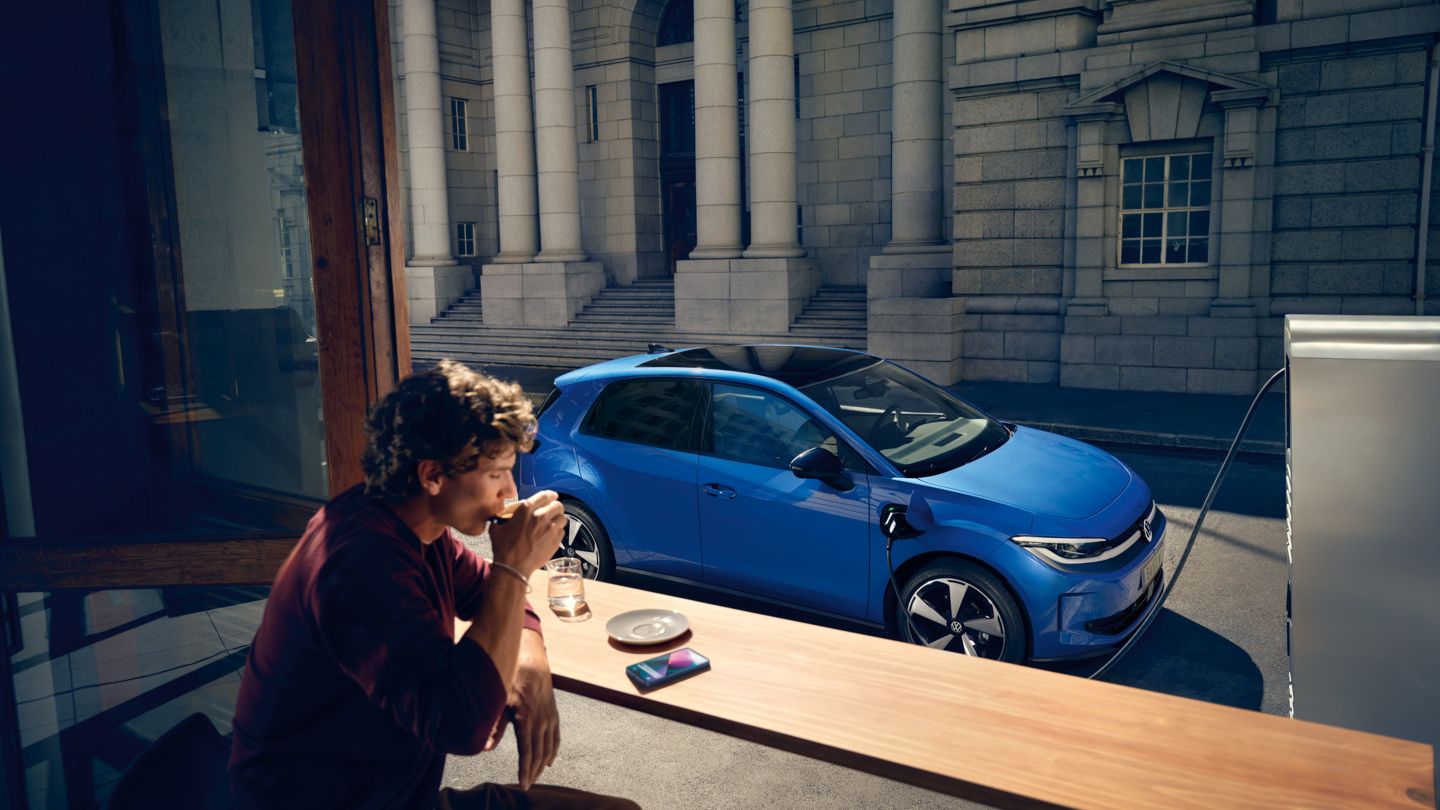 Ein Mann sitzt in einem Café, durch ein offenes Fenster ist der blaue VW ID. Polo Style zu sehen, der an einer Ladestation lädt.