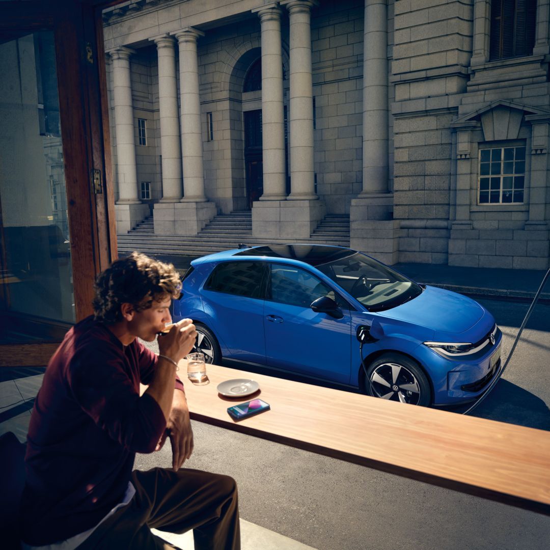 Ein Mann sitzt in einem Café, durch ein offenes Fenster ist der blaue VW ID. Polo Style zu sehen, der an einer Ladestation lädt.