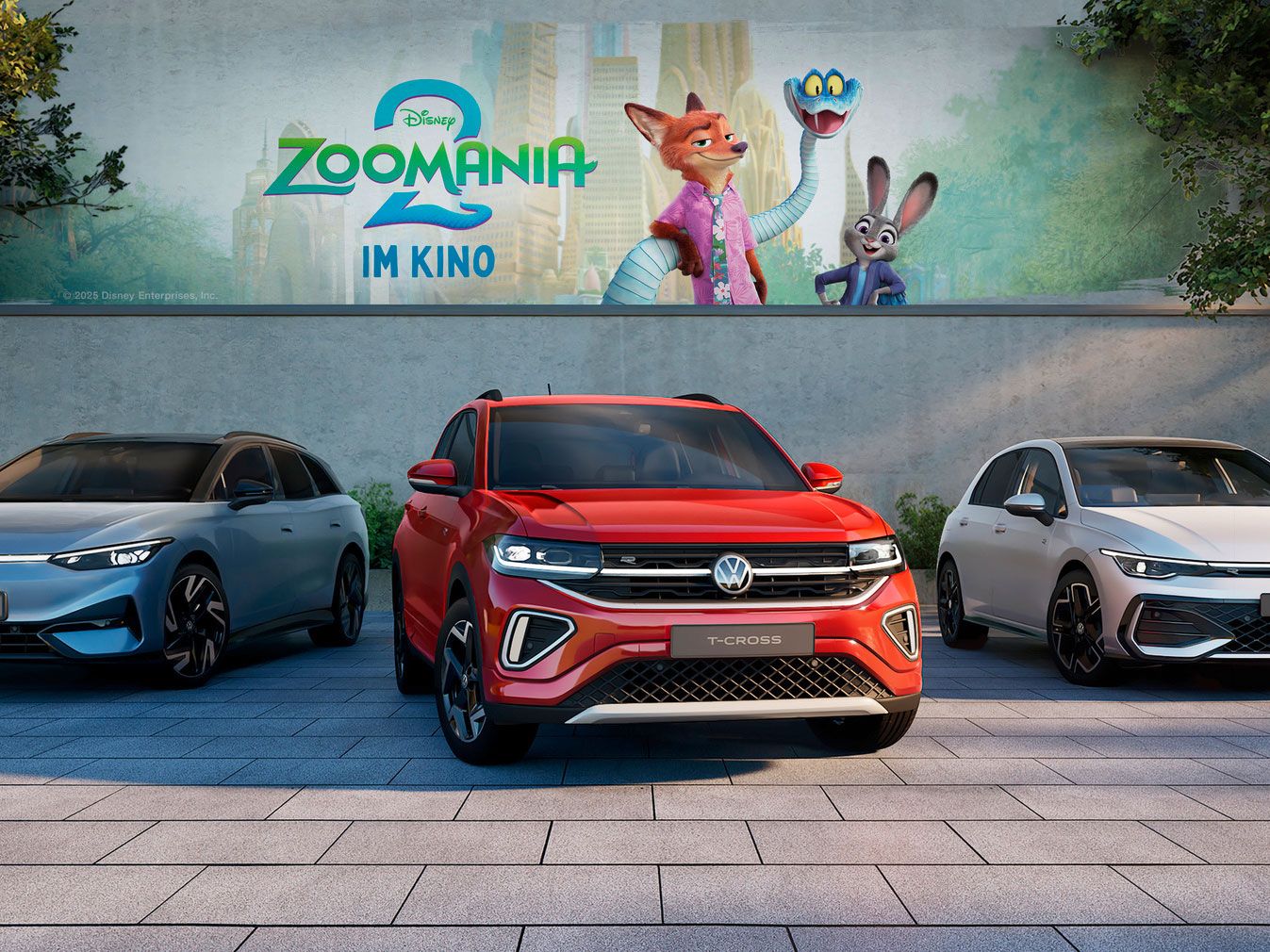 Drei Volkswagen-Fahrzeuge sind nebeneinander auf einem gepflasterten Parkplatz abgestellt. Im Hintergrund ist ein großes Plakat für den Film "Zoomania 2" zu sehen.