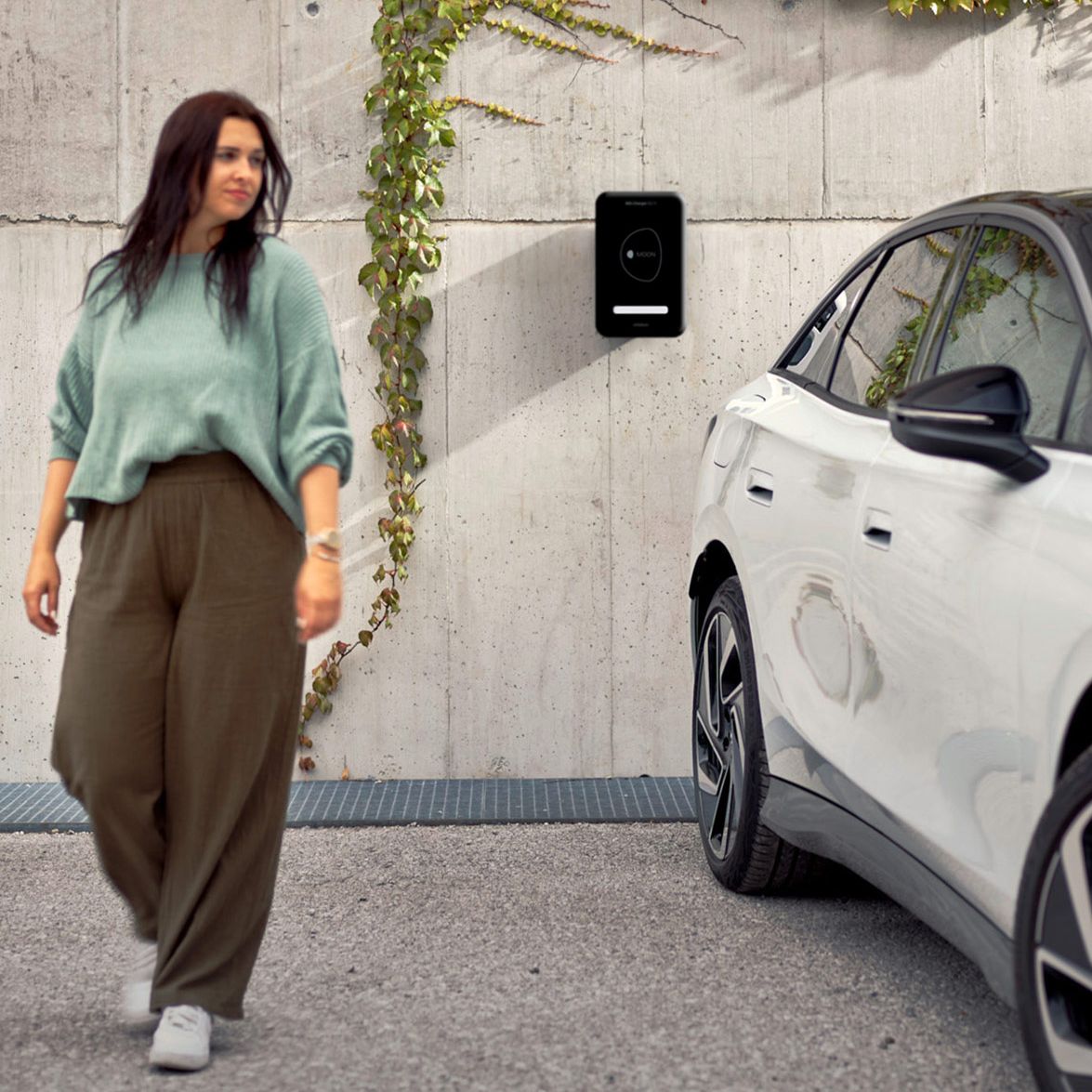 Eine Frau geht in der Nähe eines weißen ID.7, das an einer Wand mit einer Ladestation und Kletterpflanzen geparkt ist. Die Umgebung wirkt urban und modern.