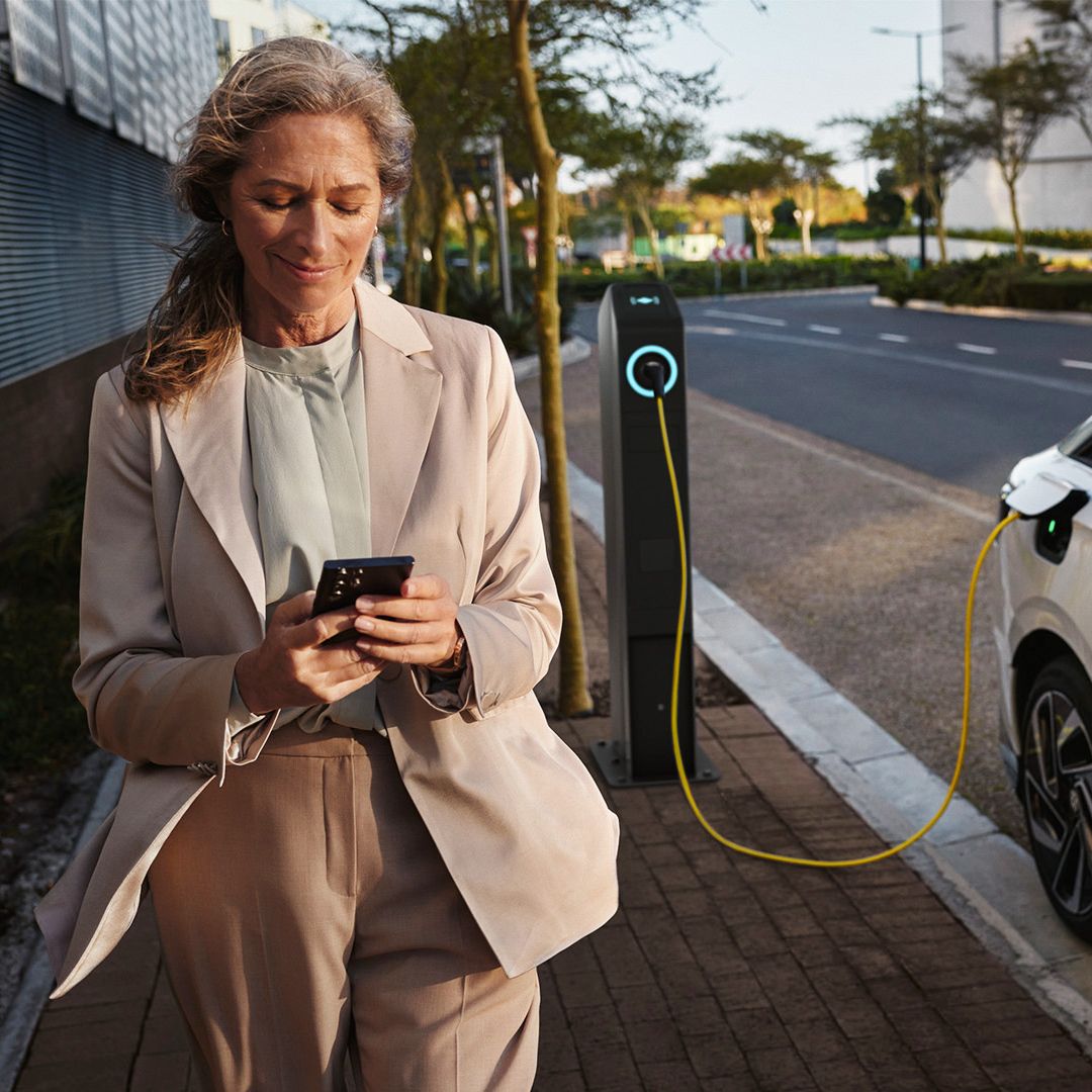 Weißer VW ID.7 seitlich lädt an einer öffentlichen Ladesäule. Eine Frau geht mit dem Smartphone in der Hand daran vorbei.