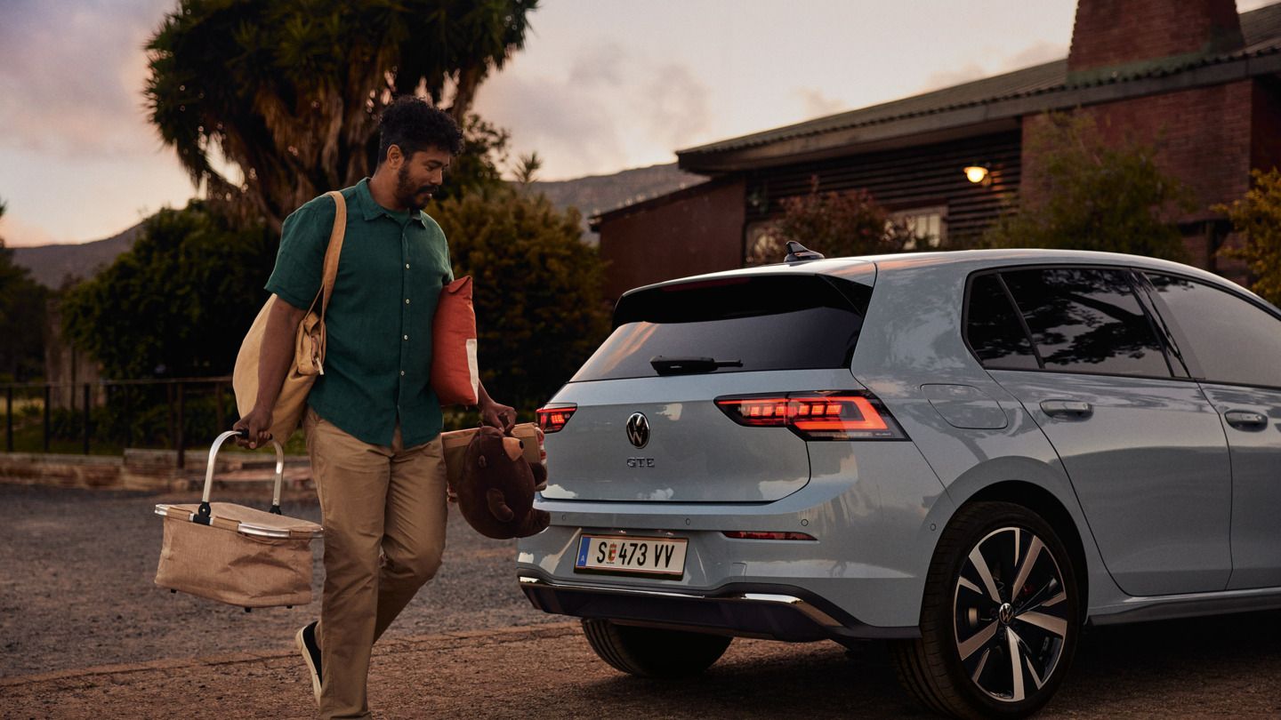 Heckansicht eines grau-blauen VW Golf GTE in abendlicher Stimmung, Mann trägt einen Korb und Spielsachen