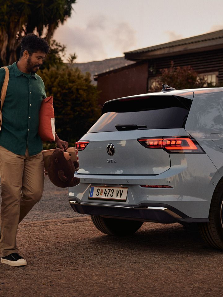 Heckansicht eines grau-blauen VW Golf GTE in abendlicher Stimmung, Mann trägt einen Korb und Spielsachen