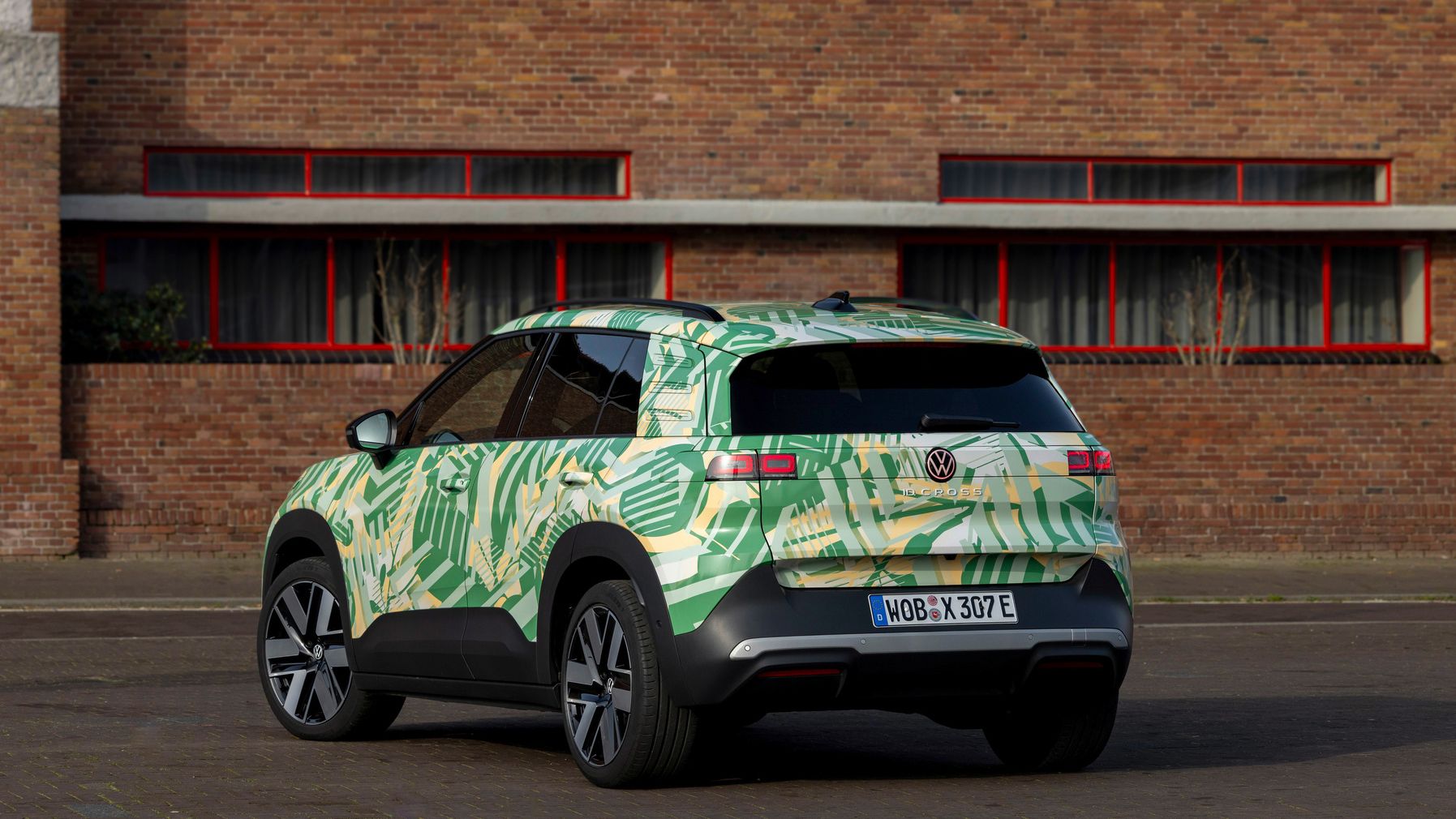 Ein Elektro-SUV der Marke Volkswagen steht auf einer gepflasterten Straße vor einer Backsteinmauer. Das Auto hat eine grüne Tarnlackierung mit Mustern und ist von hinten zu sehen. Die Fenster des Gebäudes im Hintergrund haben rote Rahmen.
