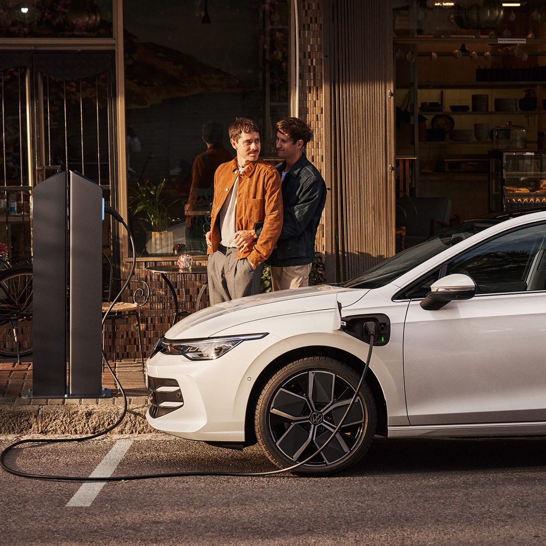 Ein weißer Golf eHybrid steht am Straßenrand und wird aufgeladen. Zwei Männer stehen neben dem Auto. 