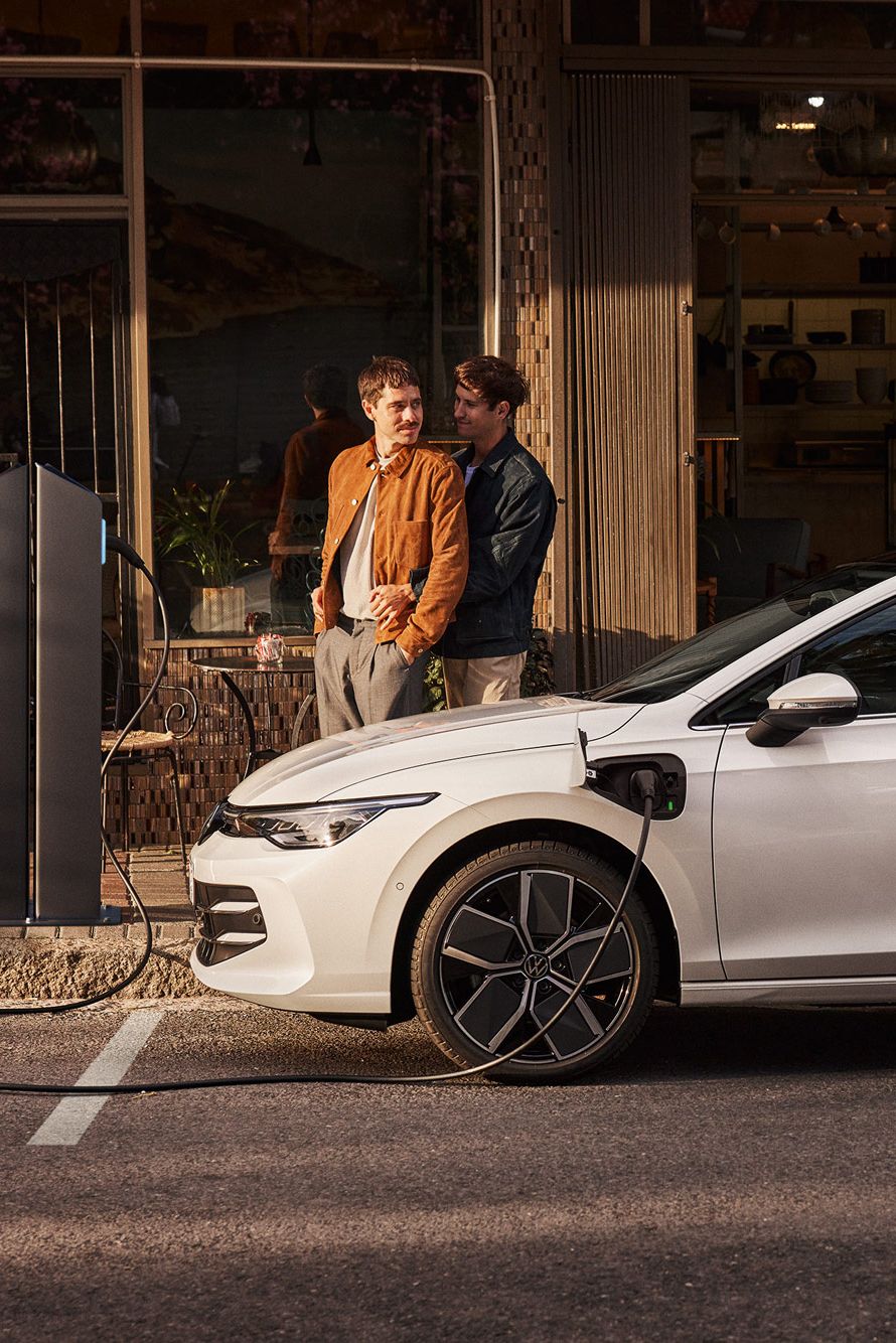 Ein weißer Golf eHybrid steht am Straßenrand und wird aufgeladen. Zwei Männer stehen neben dem Auto. 