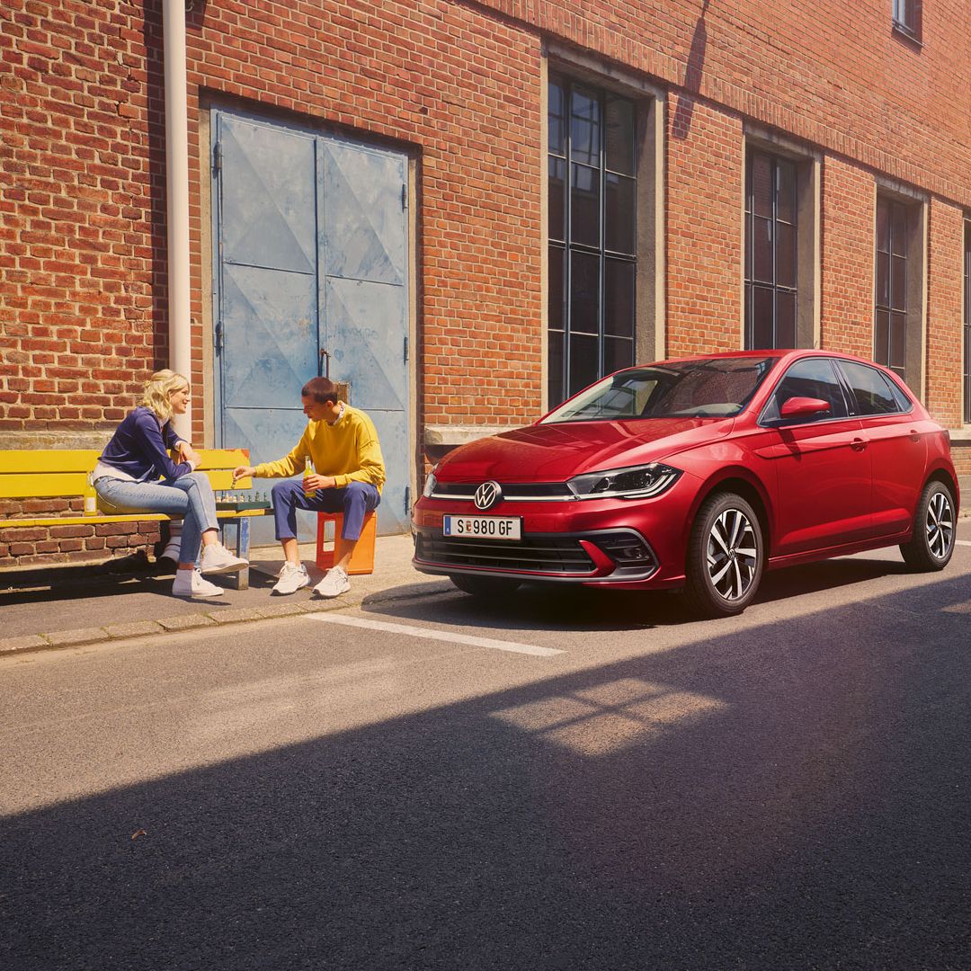 Zwei Personen spielen Schach vor einem roten VW Polo Style, der vor einem Fabrikgebäude parkt.
