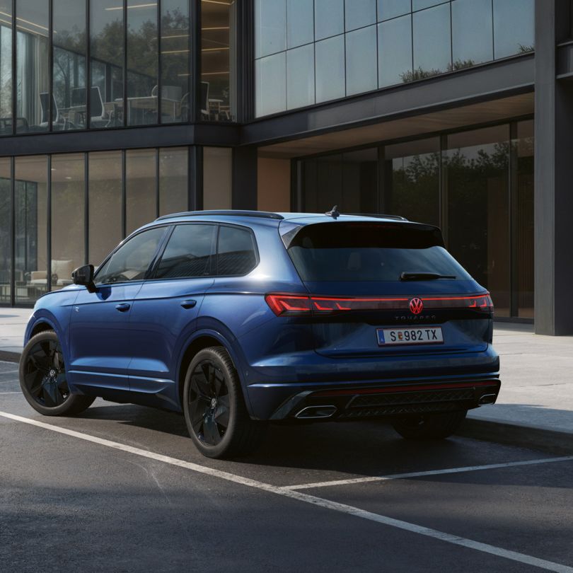Ein blauer VW Touareg FINAL EDITION  steht vor einem Bürogebäude mit Glasfassade - Ansicht von schräg hinten.
