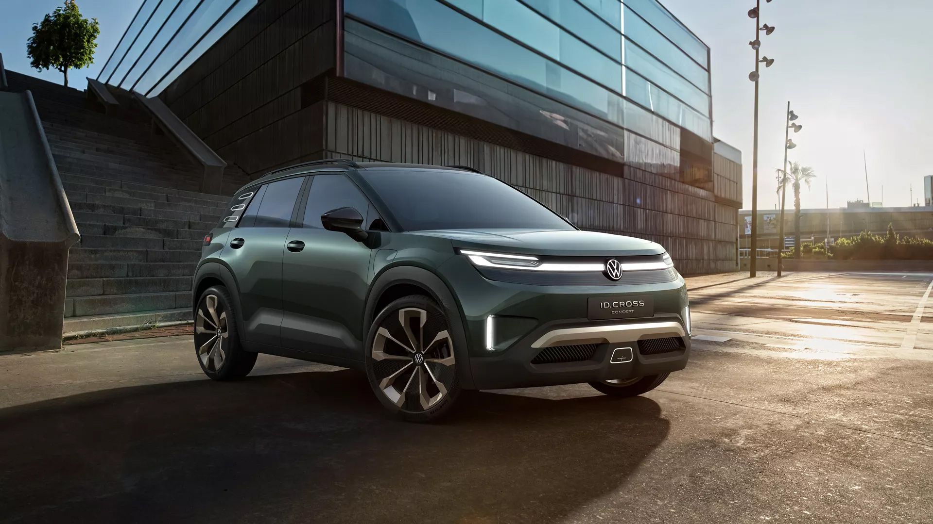 Ein dunkelgrünes Volkswagen ID. Cross steht auf einem Parkplatz vor einem modernen Gebäude mit Glasfassade. Der SUV hat futuristische Scheinwerfer und stylische Felgen. Im Hintergrund sind Treppenstufen und ein paar Palmen sichtbar.